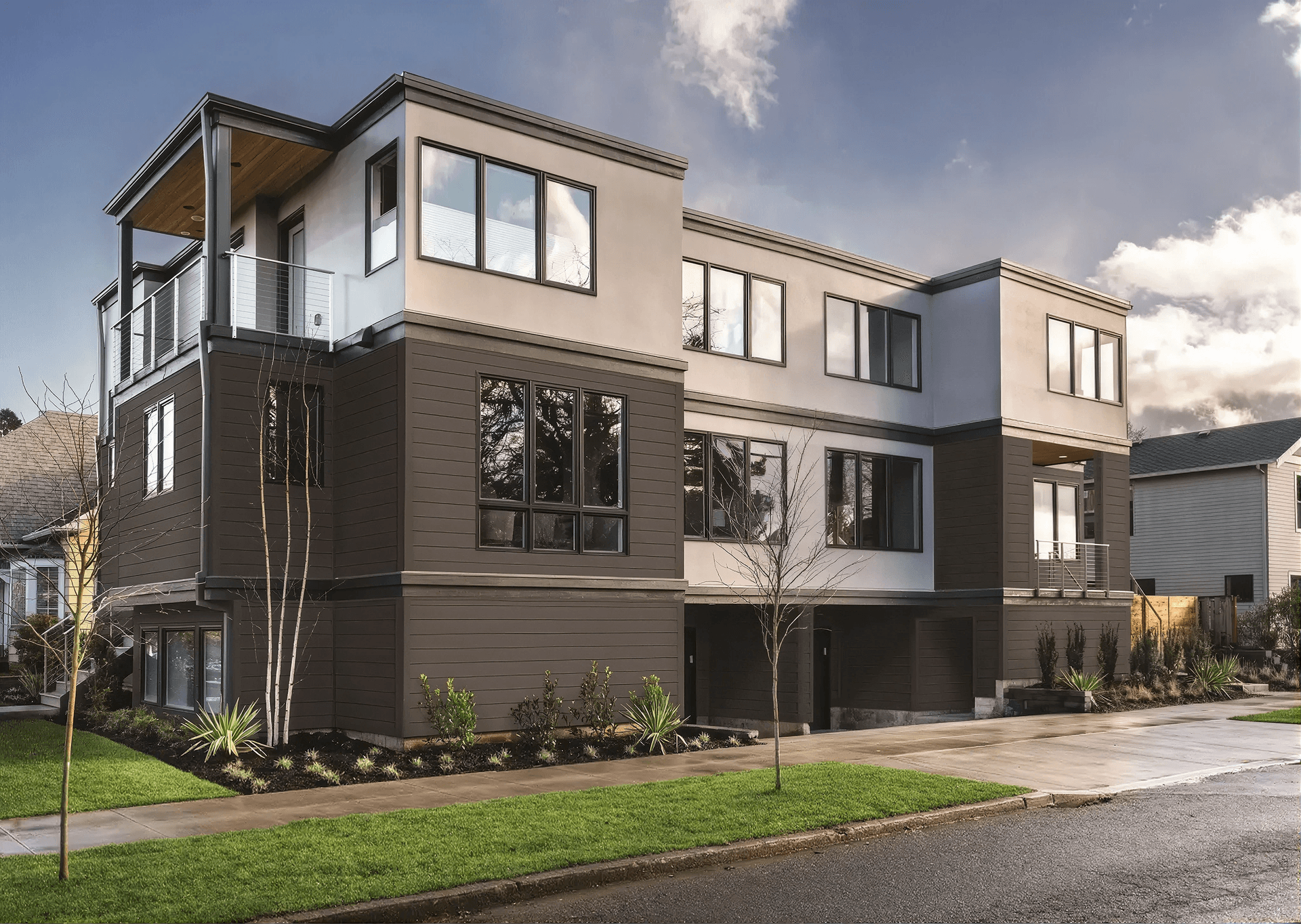 Exterior front-angle view of a modern multi-family residential building featuring dark charcoal grey horizontal composite siding on the lower levels, contrasting off-white stucco on the top floor, dark-framed multi-panel windows, a third-story balcony with metal railings, and minimalist landscaping along the sidewalk.