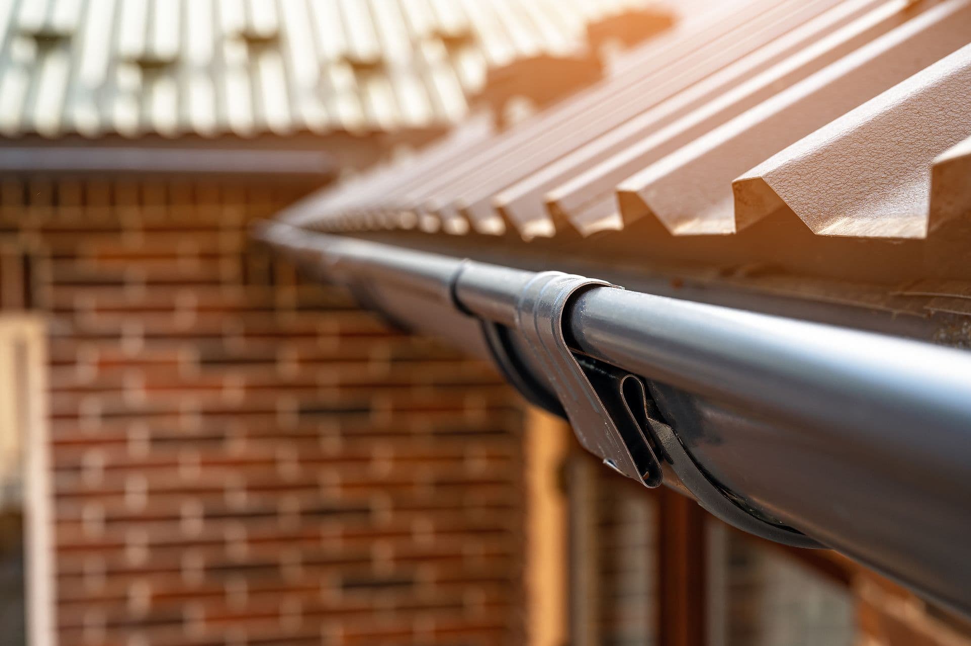 Close-up perspective shot of a dark metallic half-round gutter system attached along the edge of a corrugated metal roof on a brick building.