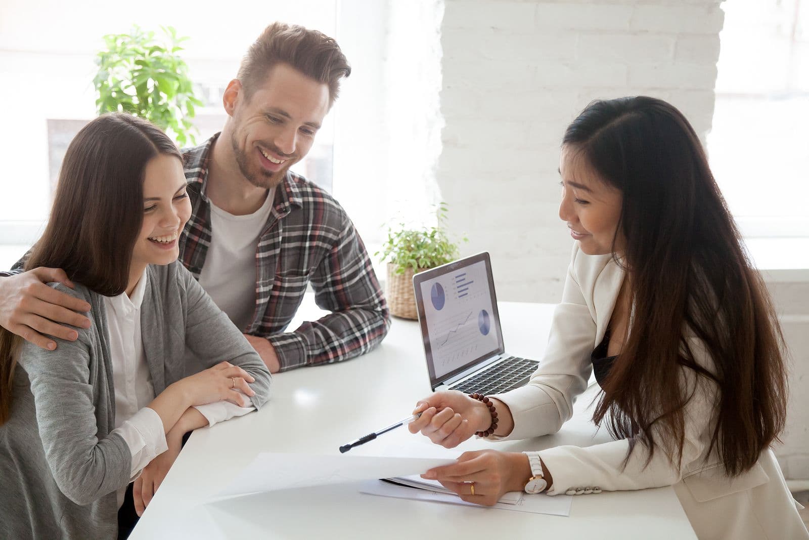 Interior view of a financial advisor or realtor in a white blazer sitting at a white desk, using a pen to point at documents for a smiling young couple seated opposite her. An open laptop showing charts and graphs rests on the table beside them in a bright, modern office setting.