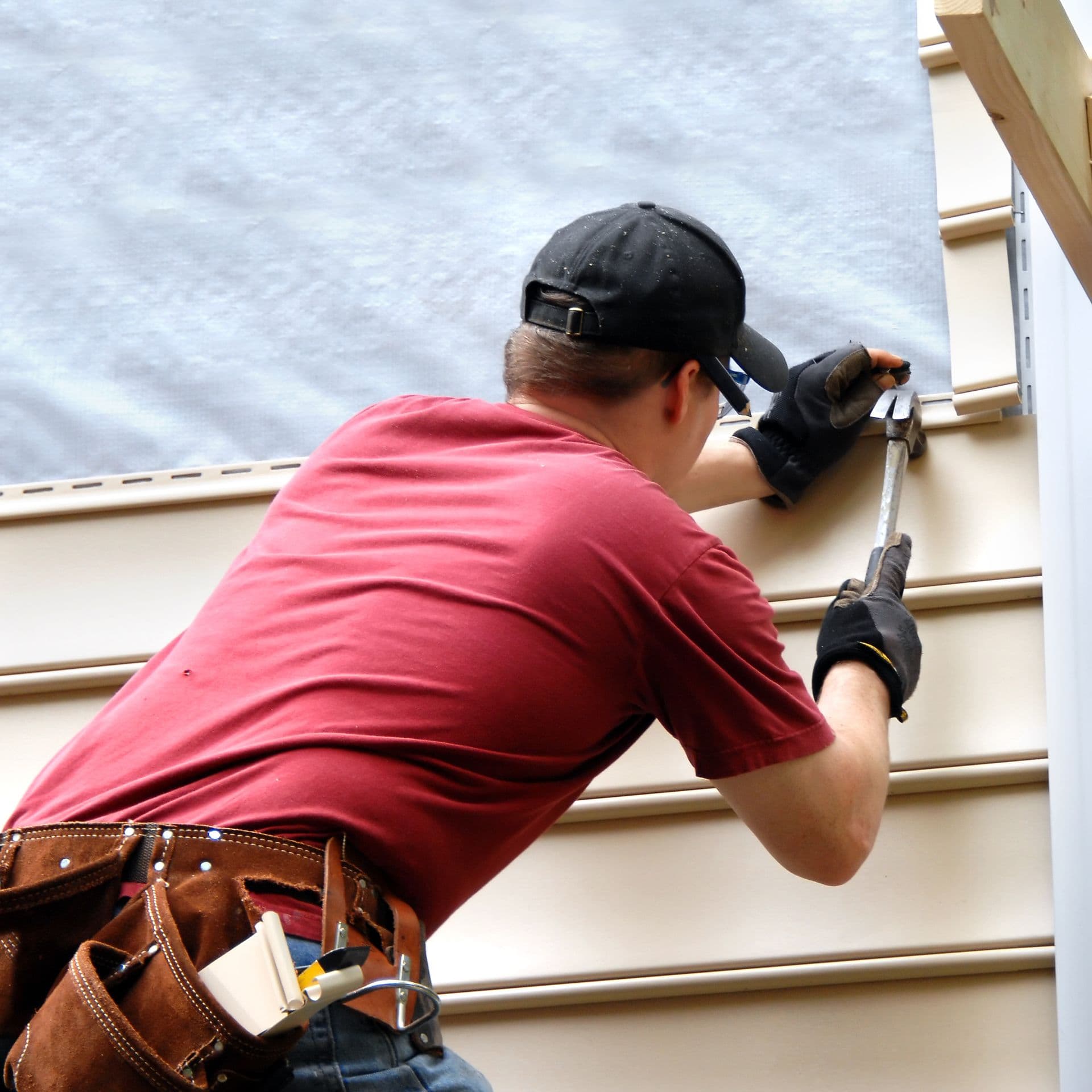 First time home buyer works to install siding on his new home. He is hammering into place a sheet of siding. He has on a red shirt and is holding hammer and nail.
