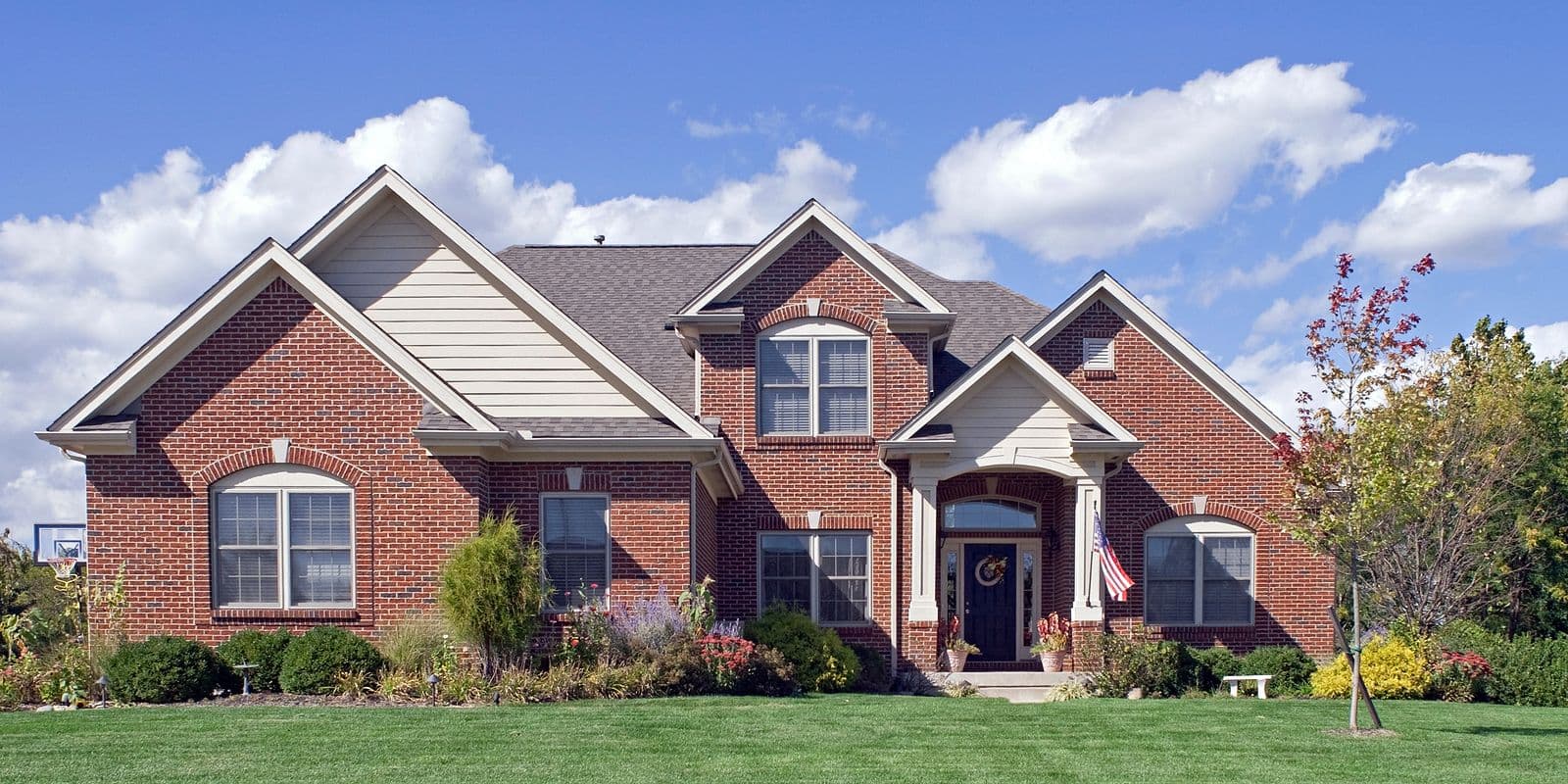 Two-story brick residential home with multiple gables, white trim, arched entry, and landscaped front yard on sunny day