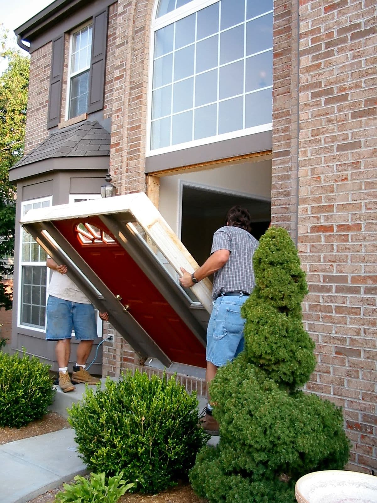 Two men removing a front door from a brick house in the suburbs.