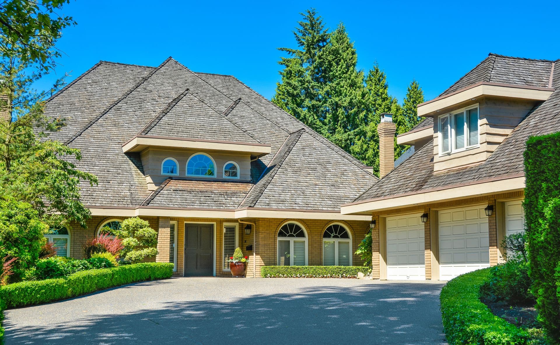 Residential house with massive roofs with green hedge at the entrance on blue sky background. Family house with big roofs tiled by wooden shingles. House with detached garage for three parking places