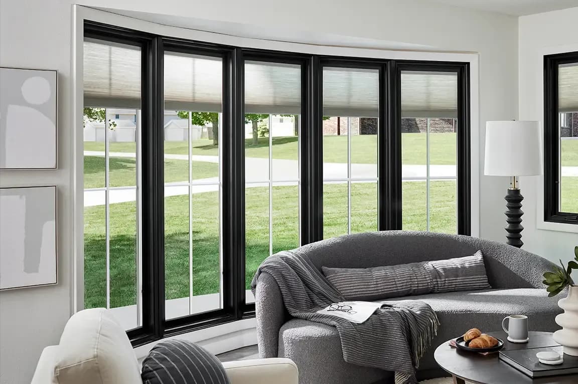 Interior view of a modern living room featuring a wide five-panel bow window with bold black frames and grid panes looking out onto a grassy suburban yard. A curved grey textured sofa with throw pillows and a blanket sits in the foreground beside a round coffee table and a standing lamp.
