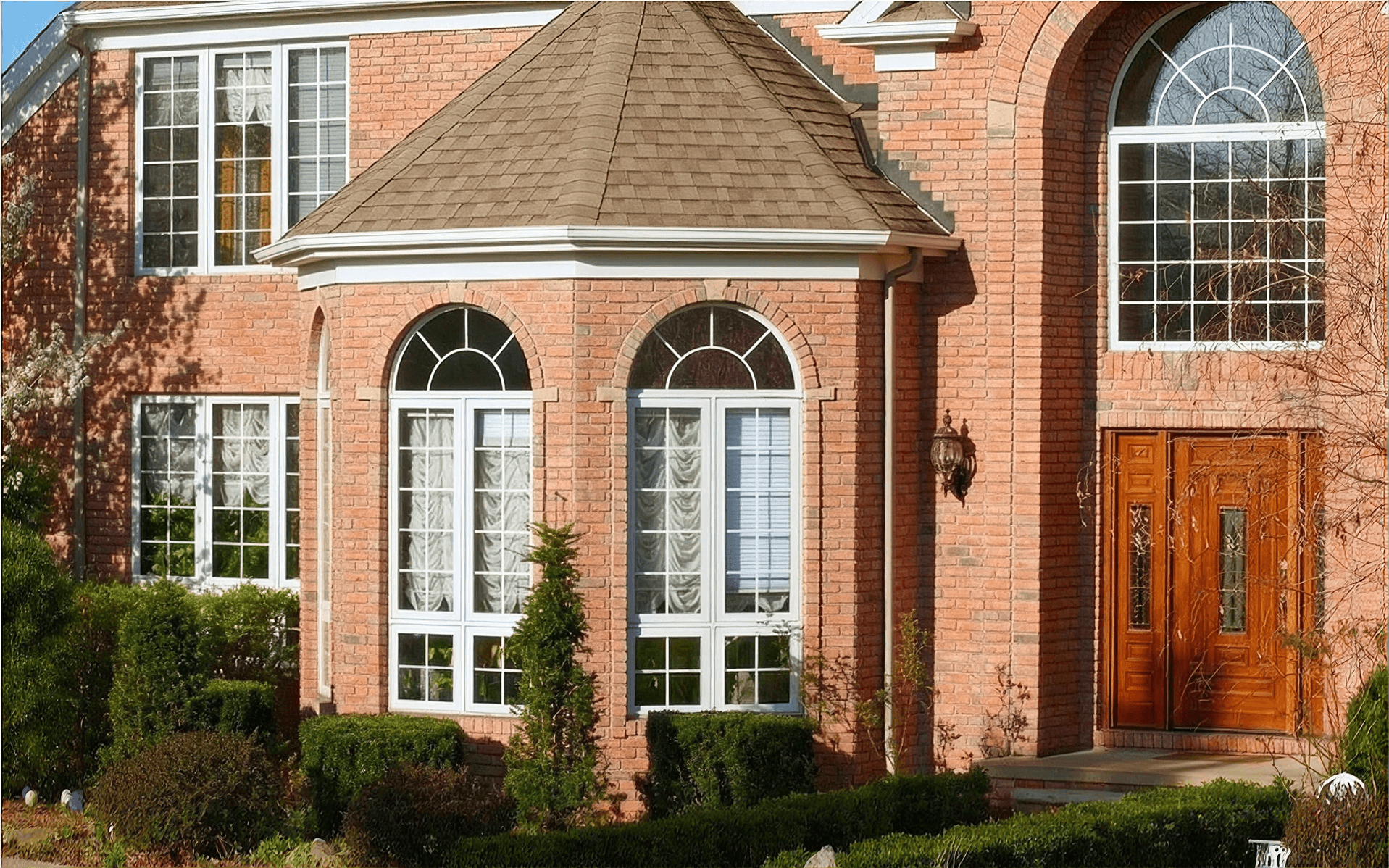 Exterior front view of a two-story red brick home featuring a two-story turret with white arched windows, decorative white grid panes, a wooden front entry door with glass side inserts, and landscaped shrubbery along the foundation.