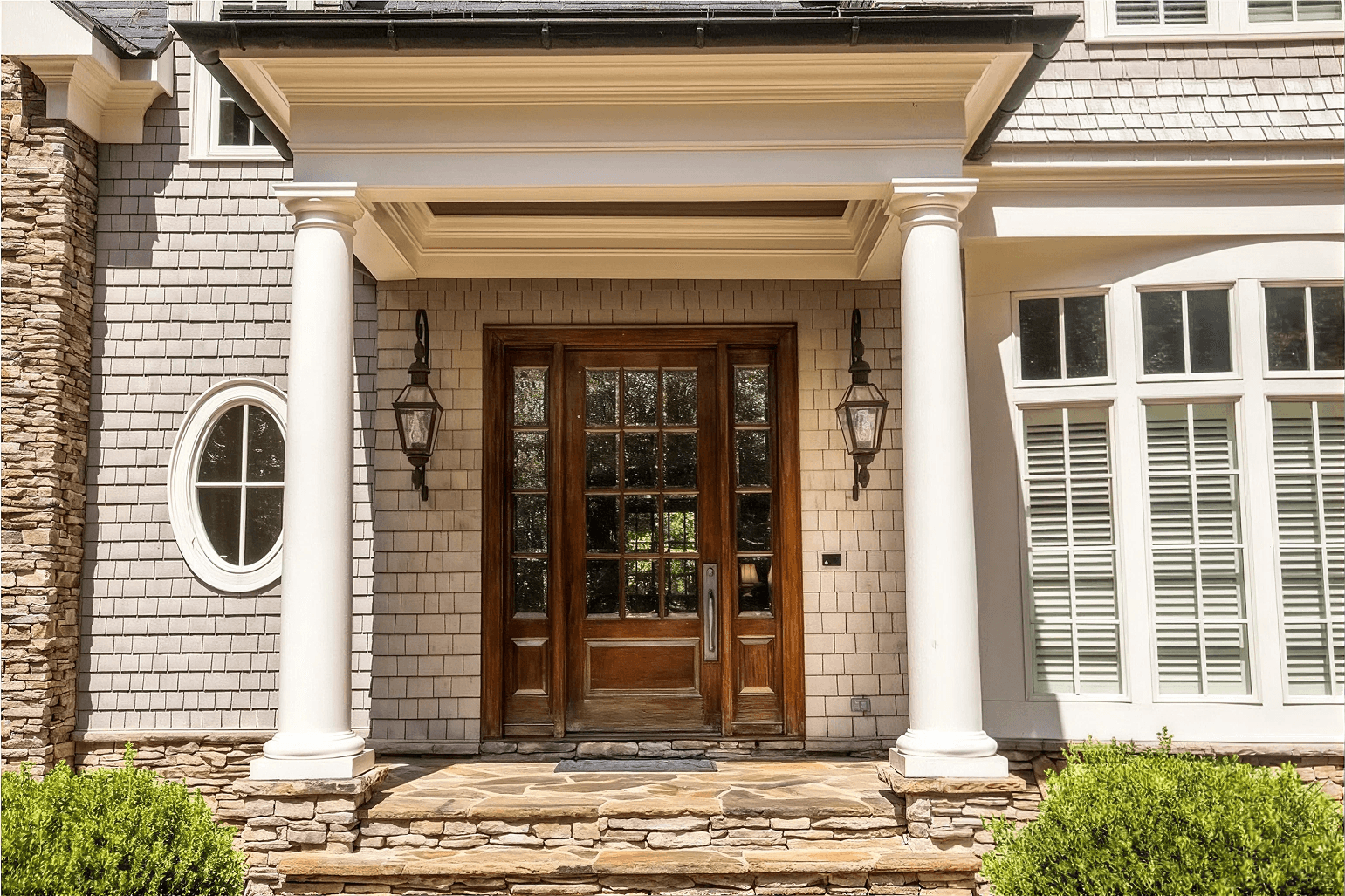 Exterior entryway view of a luxury home featuring a grand covered porch with white classical round columns and flagstone steps. The entry consists of a rich, warm wood front door with a 12-pane glass grid, flanked by matching full-length glass grid sidelights and dark hanging lantern sconces, set against light grey wood shingle siding and a small oval accent window.