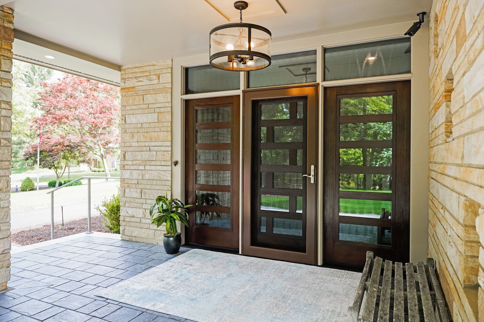 Exterior covered patio entry featuring modern, dark wood-framed double doors with horizontal glass cutouts, a central glass storm door, and transom windows above, set against light beige stone walls, slate tile flooring, a large area rug, and a rustic wooden bench.