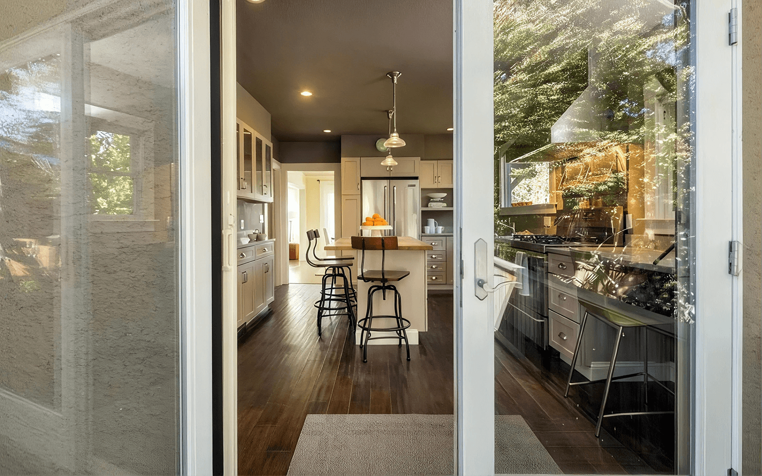 View through an open white-framed sliding glass door revealing the interior of a open-concept kitchen, featuring dark hardwood flooring, a central island with barstools, light grey cabinetry, stainless steel appliances, and pendant lighting.