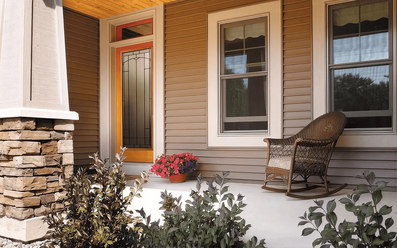 Exterior front porch view featuring an orange-trimmed front door with full-length decorative stained glass, set against brown horizontal lap siding, white-trimmed double-hung windows, a stone pillar, a wicker rocking chair, and potted pink flowers.