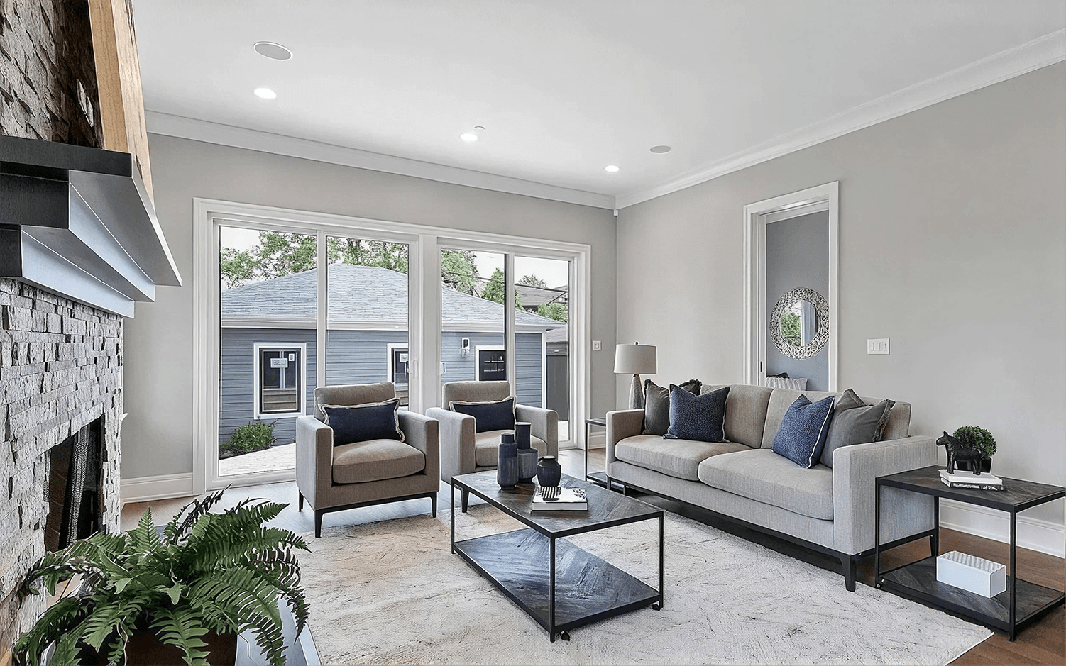 Interior living room view featuring floor-to-ceiling white-framed sliding glass patio doors that open toward an outdoor patio area, complemented by a grey sectional sofa, two accent armchairs, a stone fireplace, and modern coffee tables.