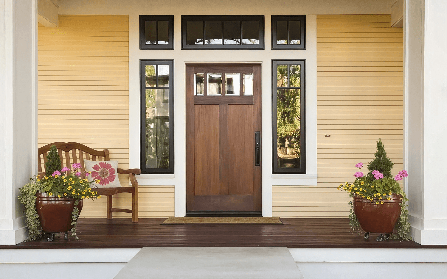 Exterior front porch view of a home with light yellow horizontal siding featuring a medium-toned wood front door with top glass grid panes, flanked by full-height black-framed sidelights and transom windows, completed with a wooden bench, potted flowers, and dark deck flooring.