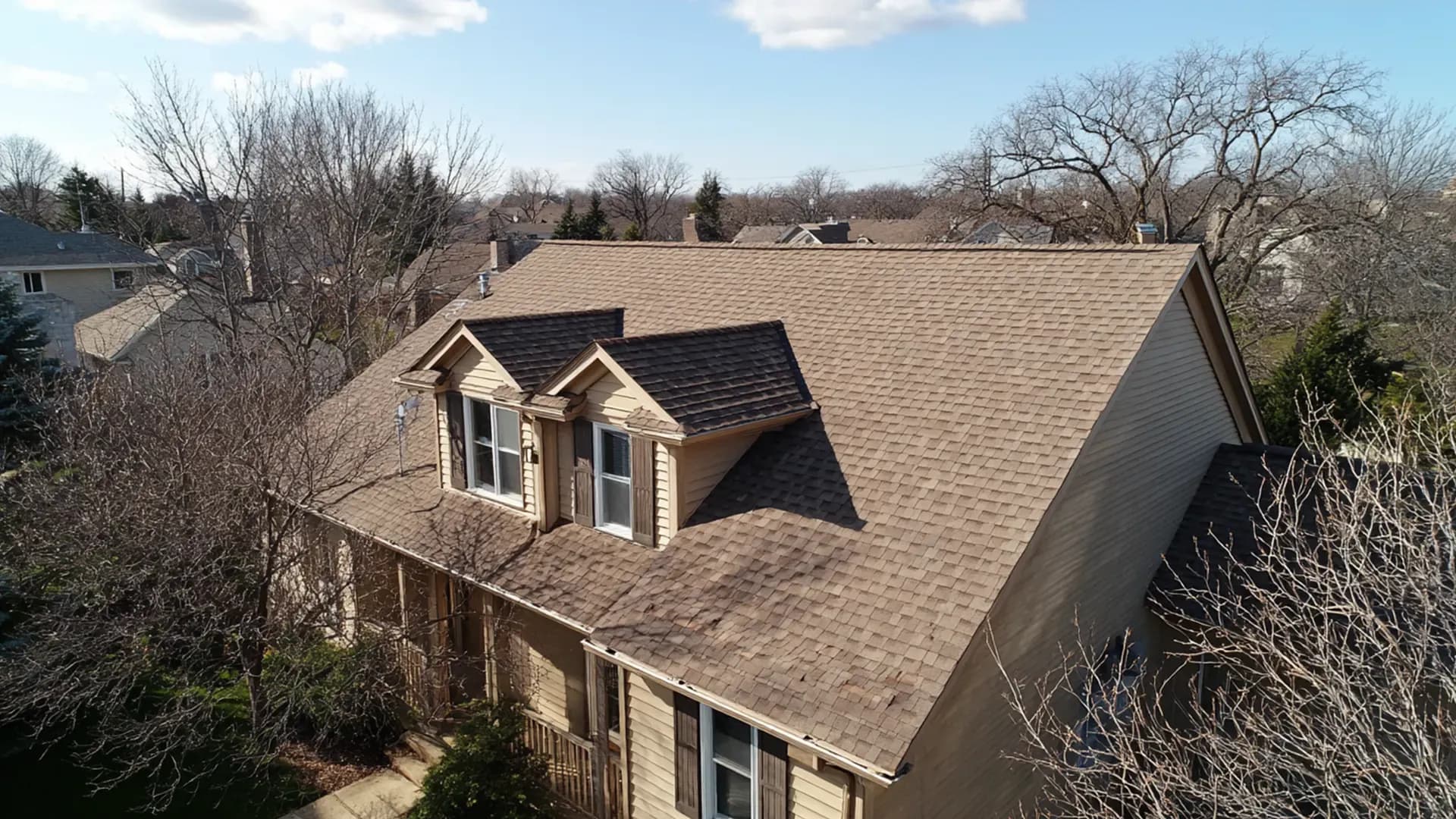 Elevated aerial view of a two-story beige home featuring a light brown architectural shingle roof, two dormer windows with shutters, and surrounding bare trees under a clear blue sky.