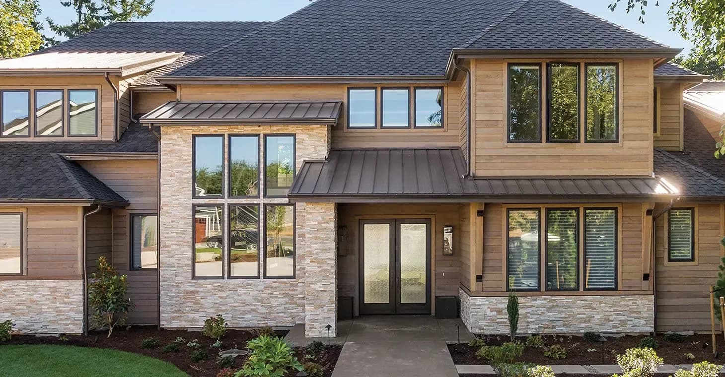 Modern two-story residential home with mixed brick and tan siding, dark metal roof, multiple windows, and dark double doors at the center entry