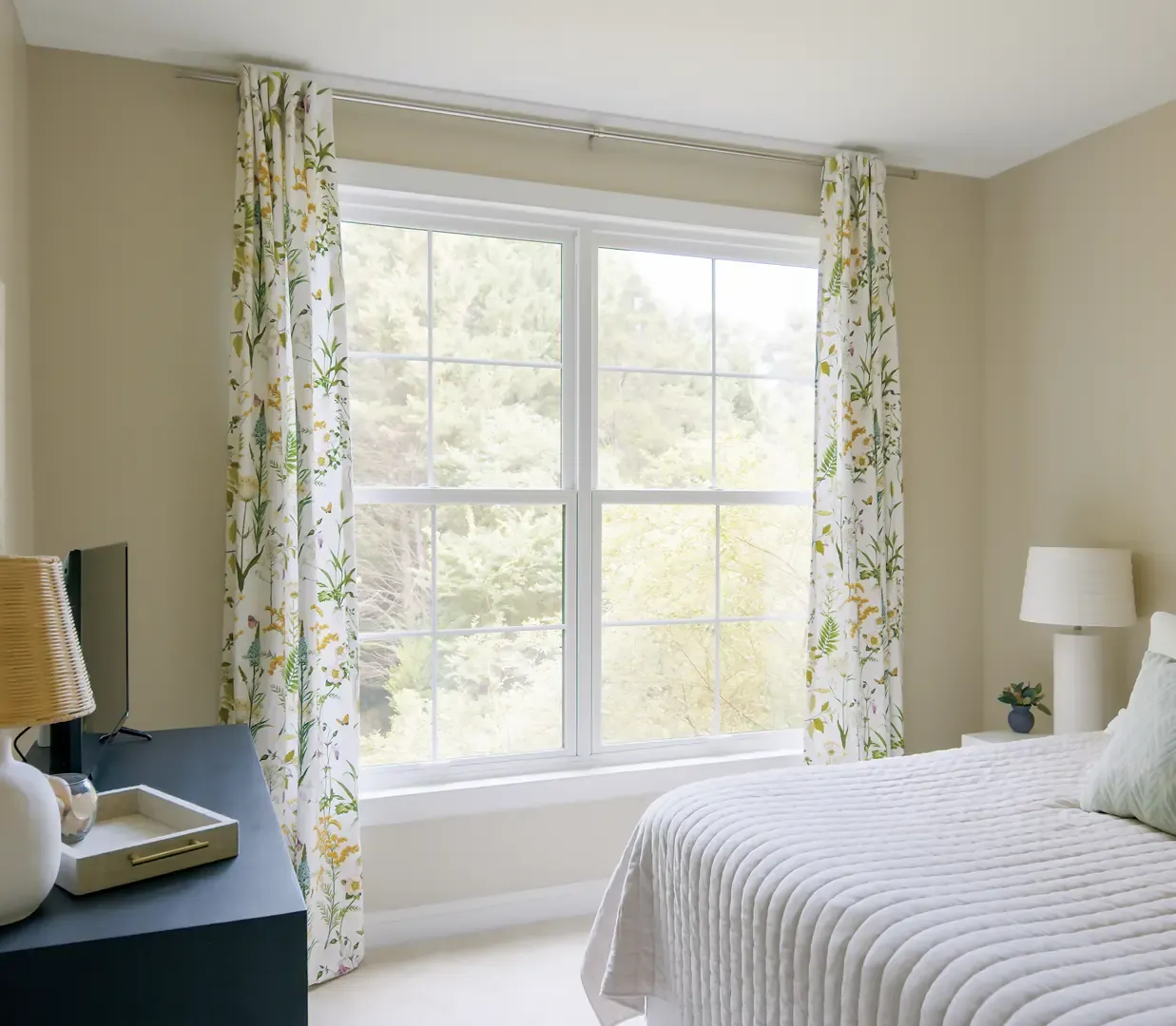 Interior view of a cozy, bright bedroom featuring a two-panel white double-hung window with grid panes framed by floral print drapes, looking out onto a wooded area. A bed with a light grey textured comforter sits in the foreground beside a nightstand and lamp, with a dark dresser on the left.