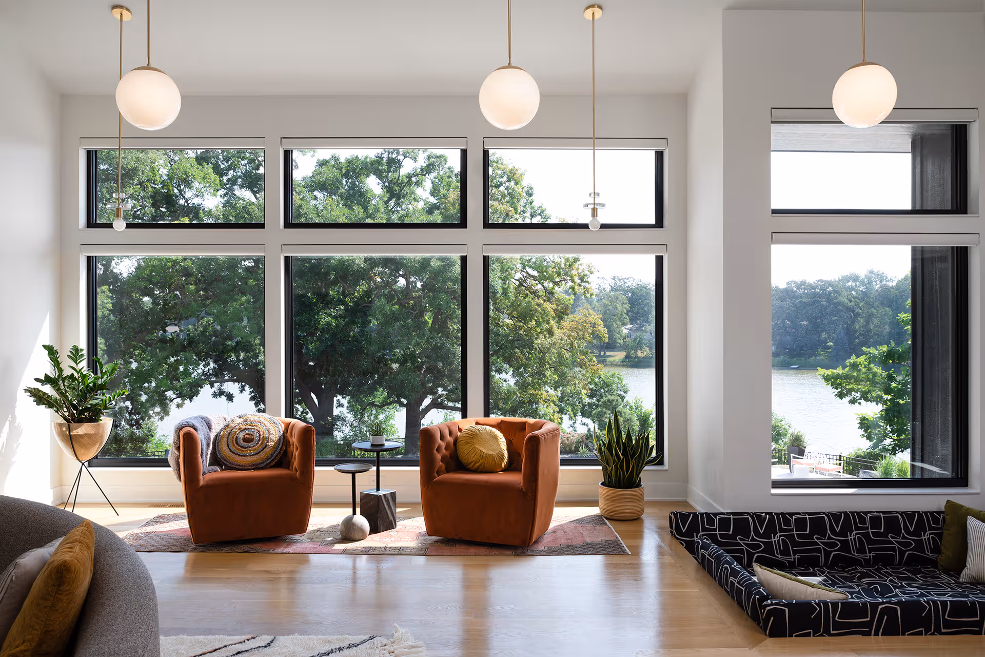 Interior view of a modern living area with floor-to-ceiling black picture windows looking out onto a lake and green trees, furnished with two burnt-orange velvet armchairs, pendant globe lights, potted house plants, and a patterned dog bed.