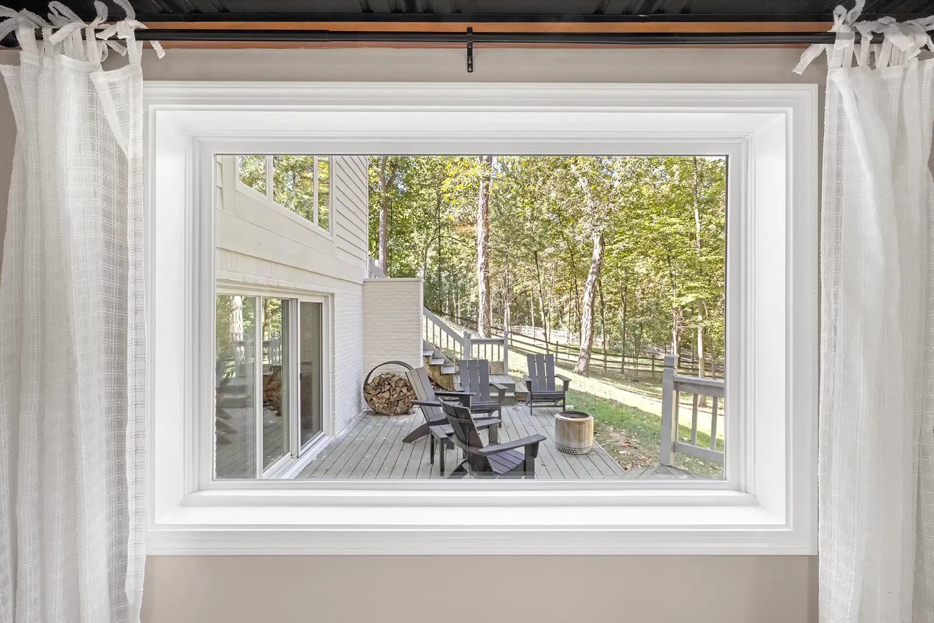Interior view looking through a white-framed picture window styled with shear white sheer drapes, framing an outdoor wooden back deck with Adirondack chairs, a fire pit, stack of firewood, and a surrounding wooded backyard.