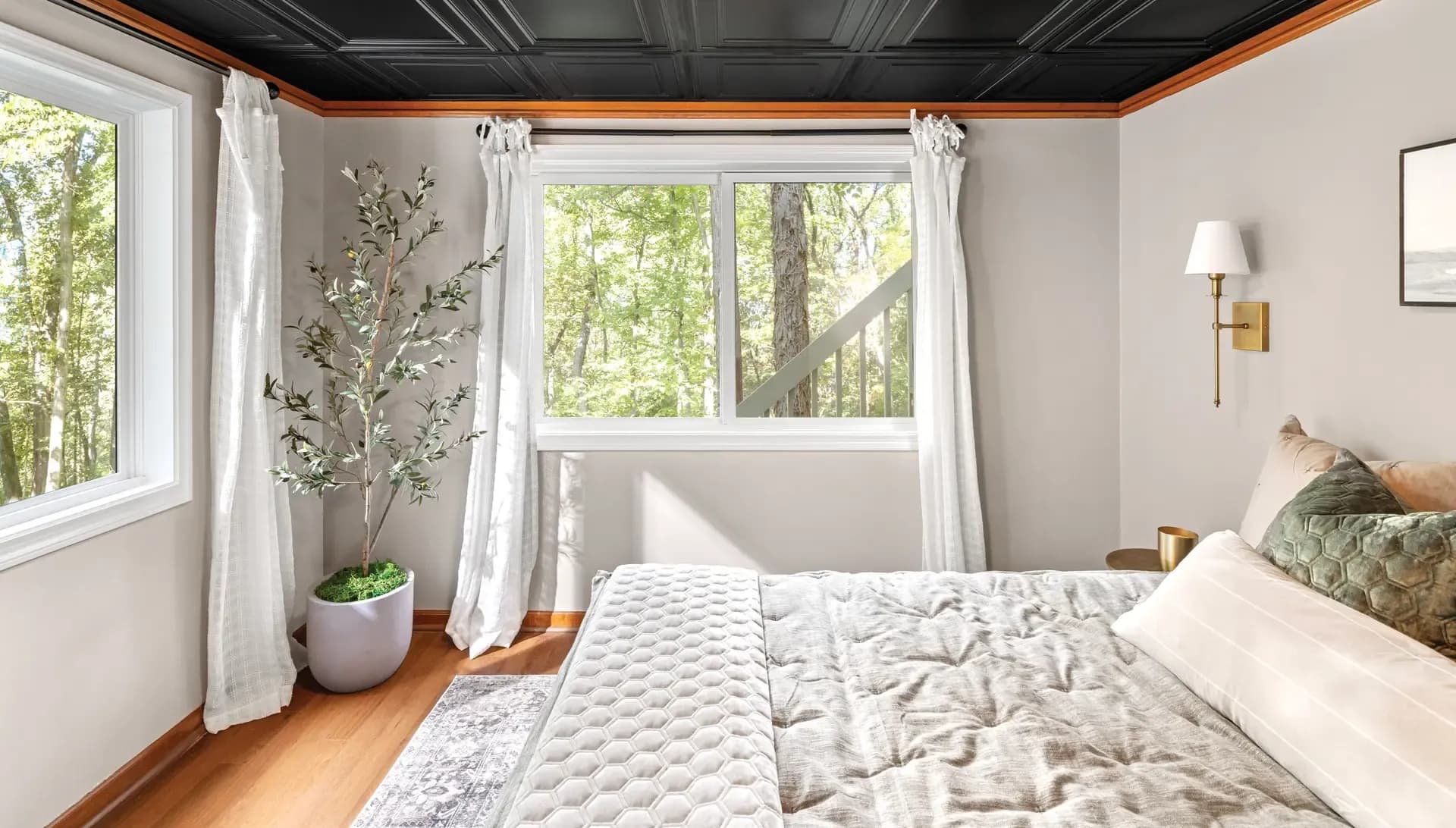 Interior view of a cozy bedroom featuring a white horizontal sliding window framed by sheer white curtains, looking out toward a wooded outdoor area. The room includes a black wainscoted ceiling, a pot plant in the corner, and a bed with a textured grey comforter.