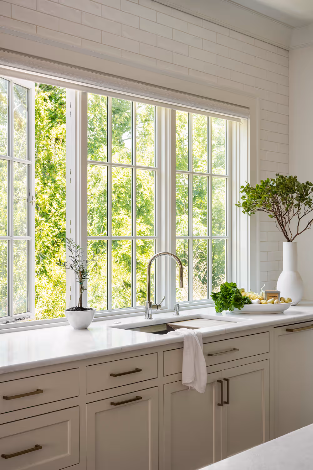 Interior kitchen view featuring a three-panel white grid window set above a marble countertop and sink, with the leftmost casement pane cranked wide open toward a bright green tree canopy outside.