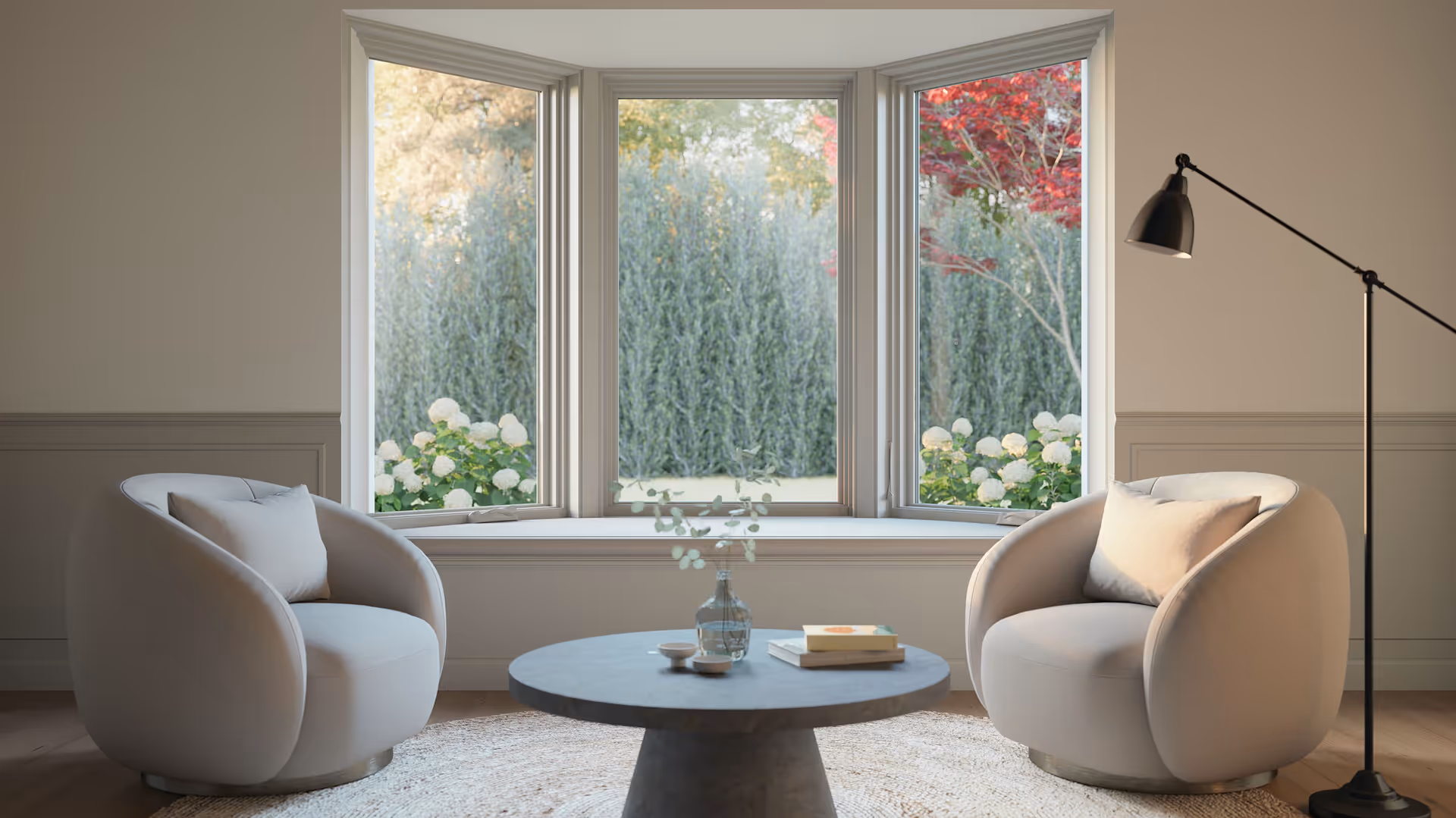 Interior view of an elegant sitting room featuring a three-panel bay window looking out onto a garden with white hydrangeas and red autumn foliage. Two round, light grey armchairs flank a low circular coffee table set on a patterned area rug, lit by a black floor lamp.