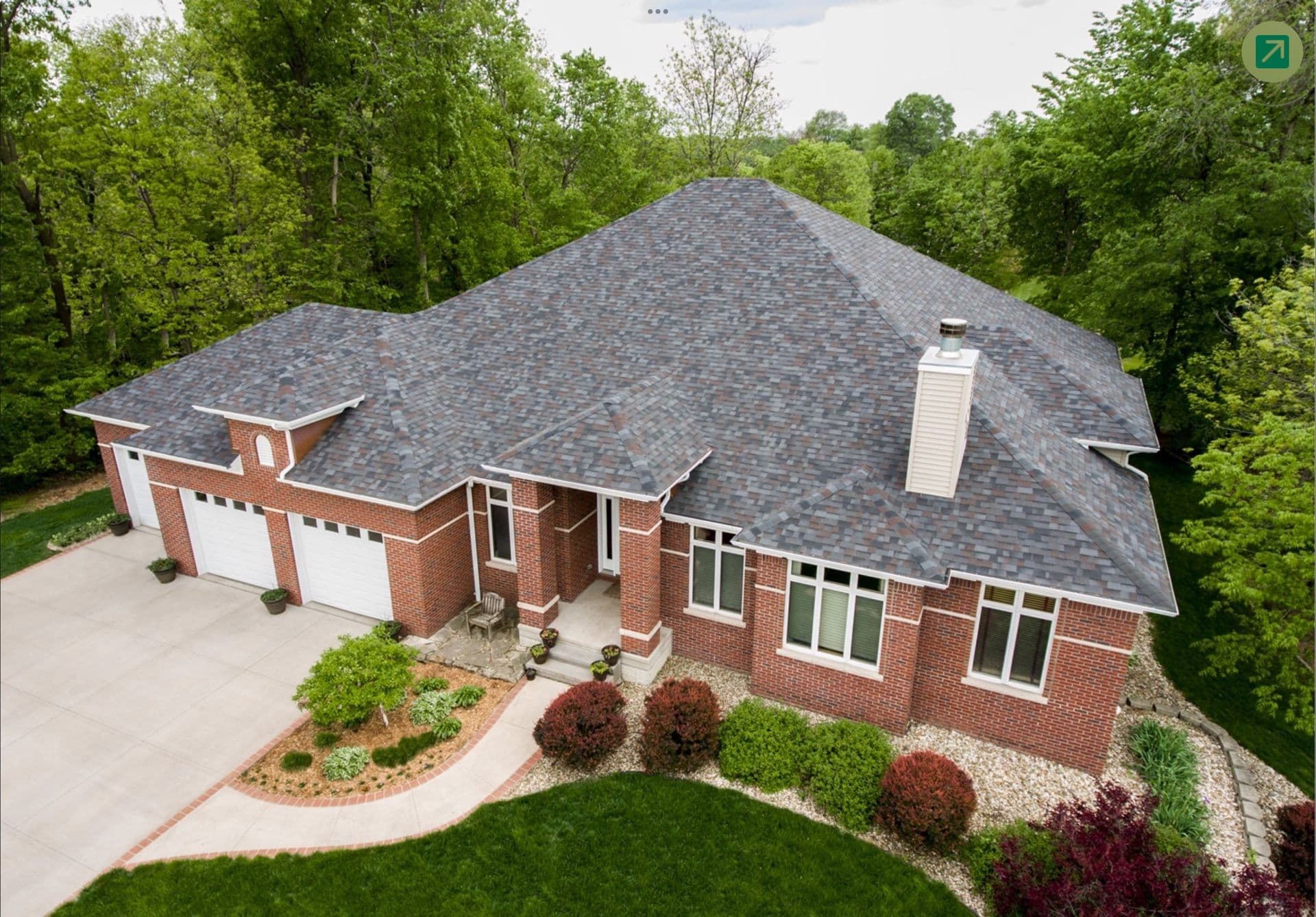 Aerial view of a brick residential home with gray asphalt shingle roof, two-car garage, front entrance with columns, and landscaped yard surrounded by trees