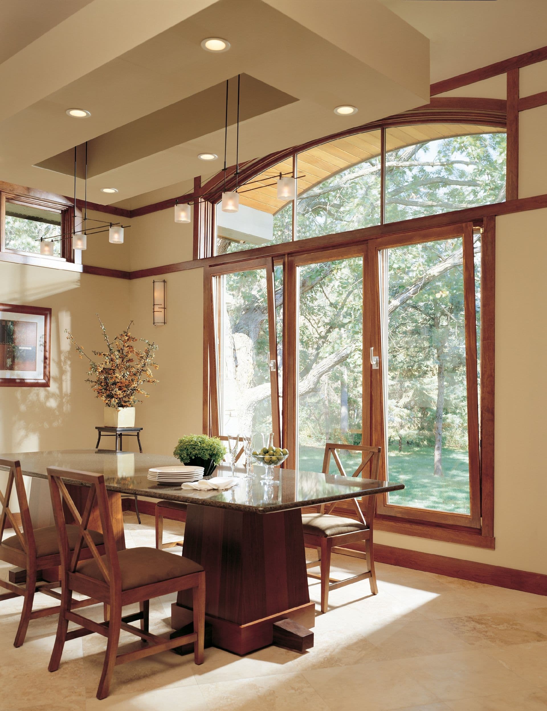 Interior dining room view featuring floor-to-ceiling, wood-framed tilt-and-turn glass doors topped by a large arched transom window looking out onto a wooded yard. A dark wood dining table with matching chairs sits on a light tiled floor beneath a tray ceiling with pendant lighting.