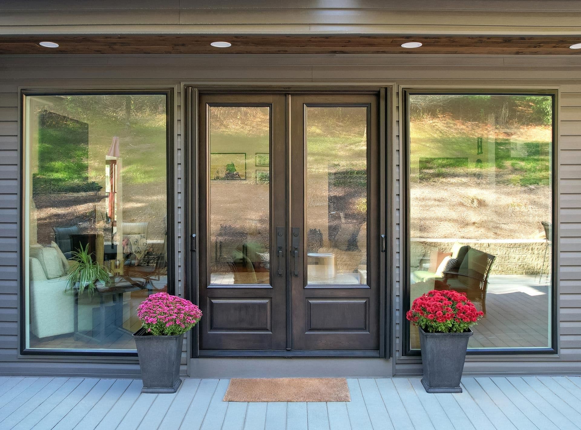 Exterior patio view featuring 8-foot dark Coffee Bean-stained cherry French doors with glass panels, flanked by large Coal Black Endure picture windows that reflect the outdoor landscape while revealing the interior seating area. The entryway is finished with grey composite decking, a doormat, and potted bright pink and red flowers.