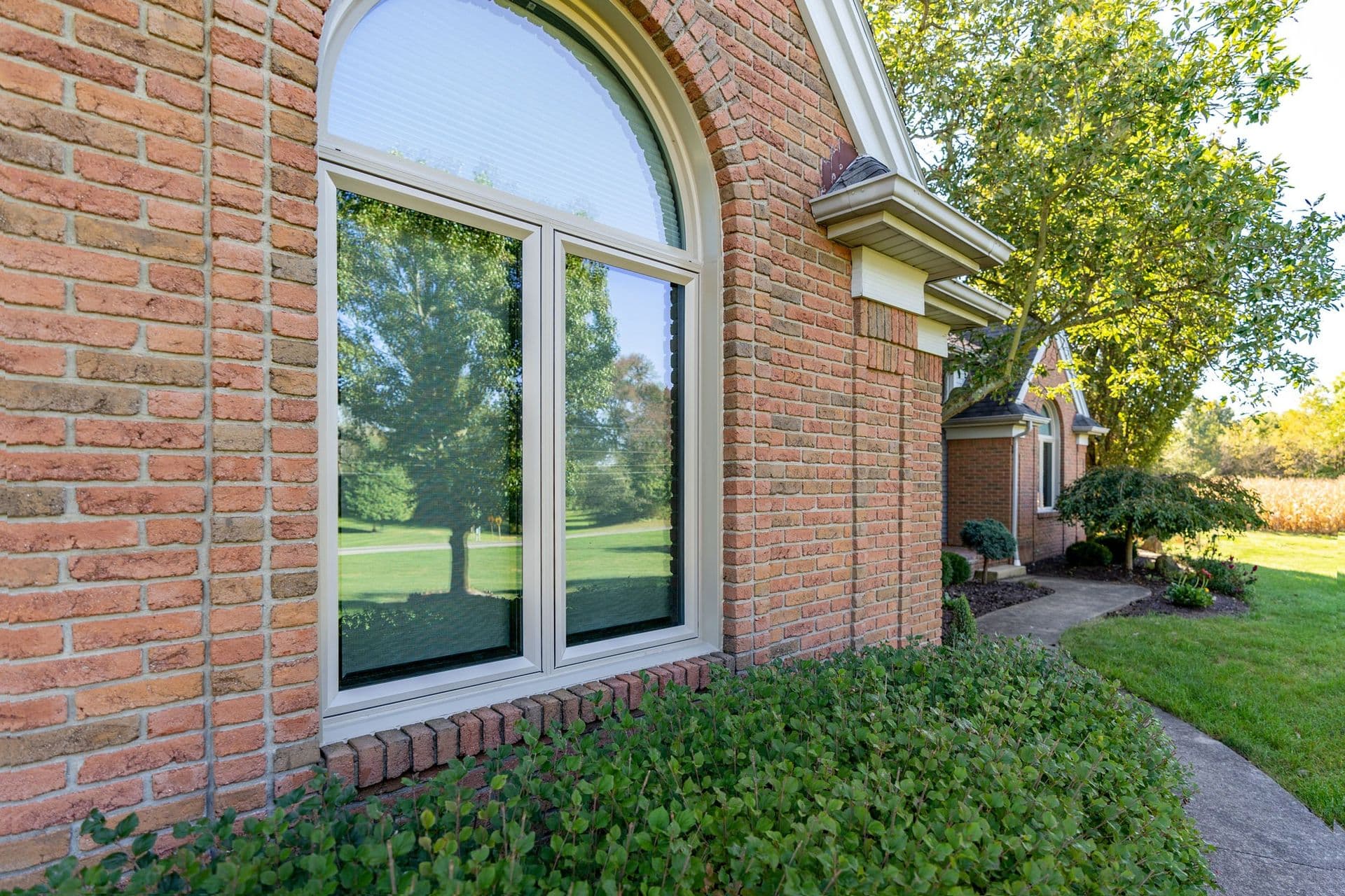 Exterior view of a sandstone-colored two-panel casement window topped by a half-round arched transom window, set in a red-and-brown brick home facade above a low green shrub garden and paved walkway.