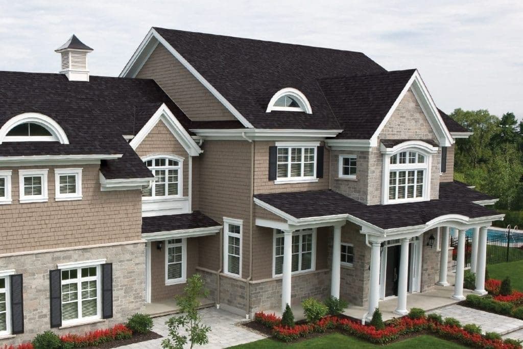 Elevated exterior view of a large, luxurious two-story home featuring tan cedar-shake siding, light-colored stone veneer lower walls, a dark grey or black shingled roof with small eyebrow dormers, and white-columned porticos framing the entrance.
