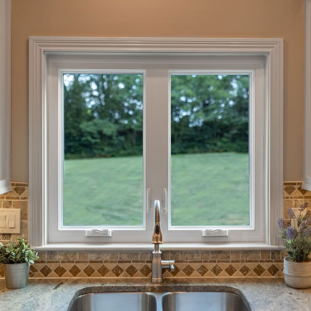 Interior view of a two-panel white casement window installed above a kitchen sink with a brushed nickel faucet, set against warm tan walls and a decorative stone backsplash.