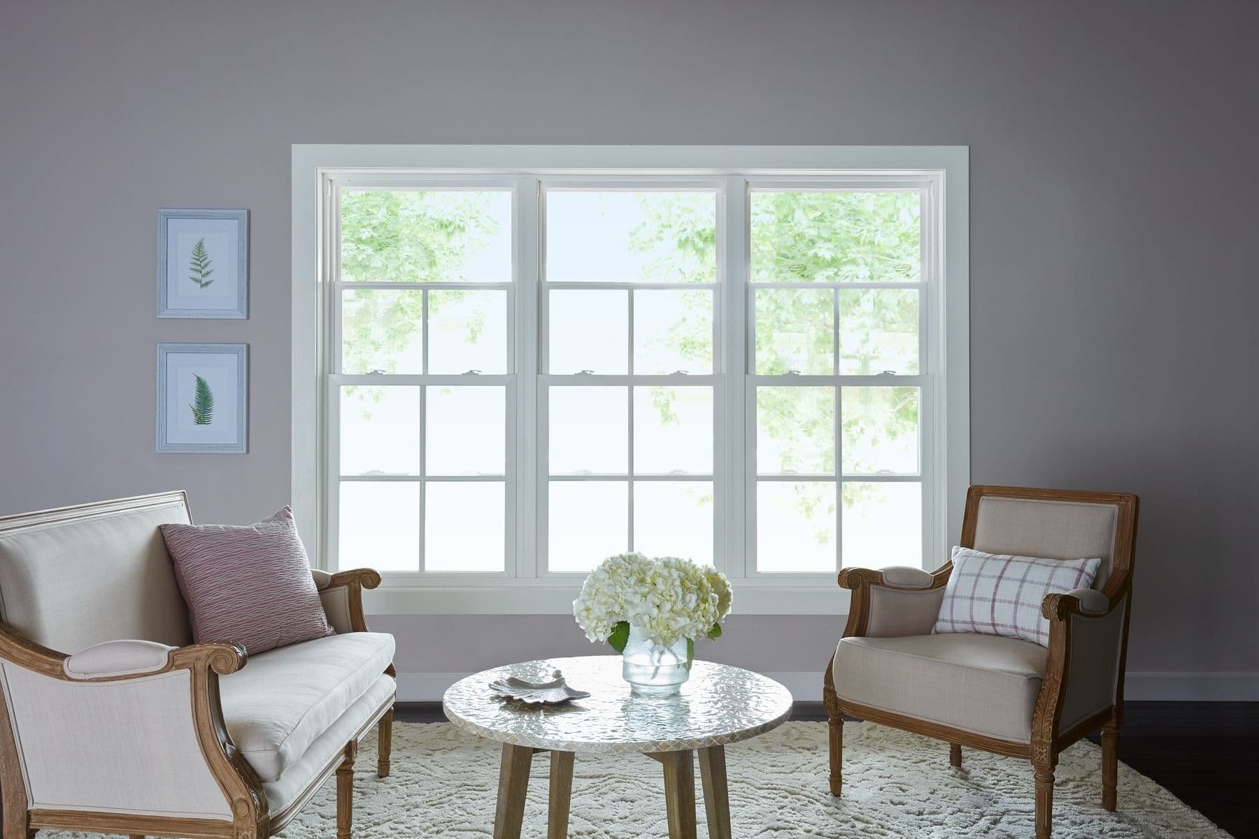 imgi_1_Double-Hung-Main.pg_.jpg Interior view of a elegant sitting room with soft grey walls, featuring three large white double-hung windows with grilles side by side. The room is decorated with a cream love seat, a matching armchair with wooden frames, a textured white coffee table with fresh flowers, and two small framed leaf prints on the wall to the left.