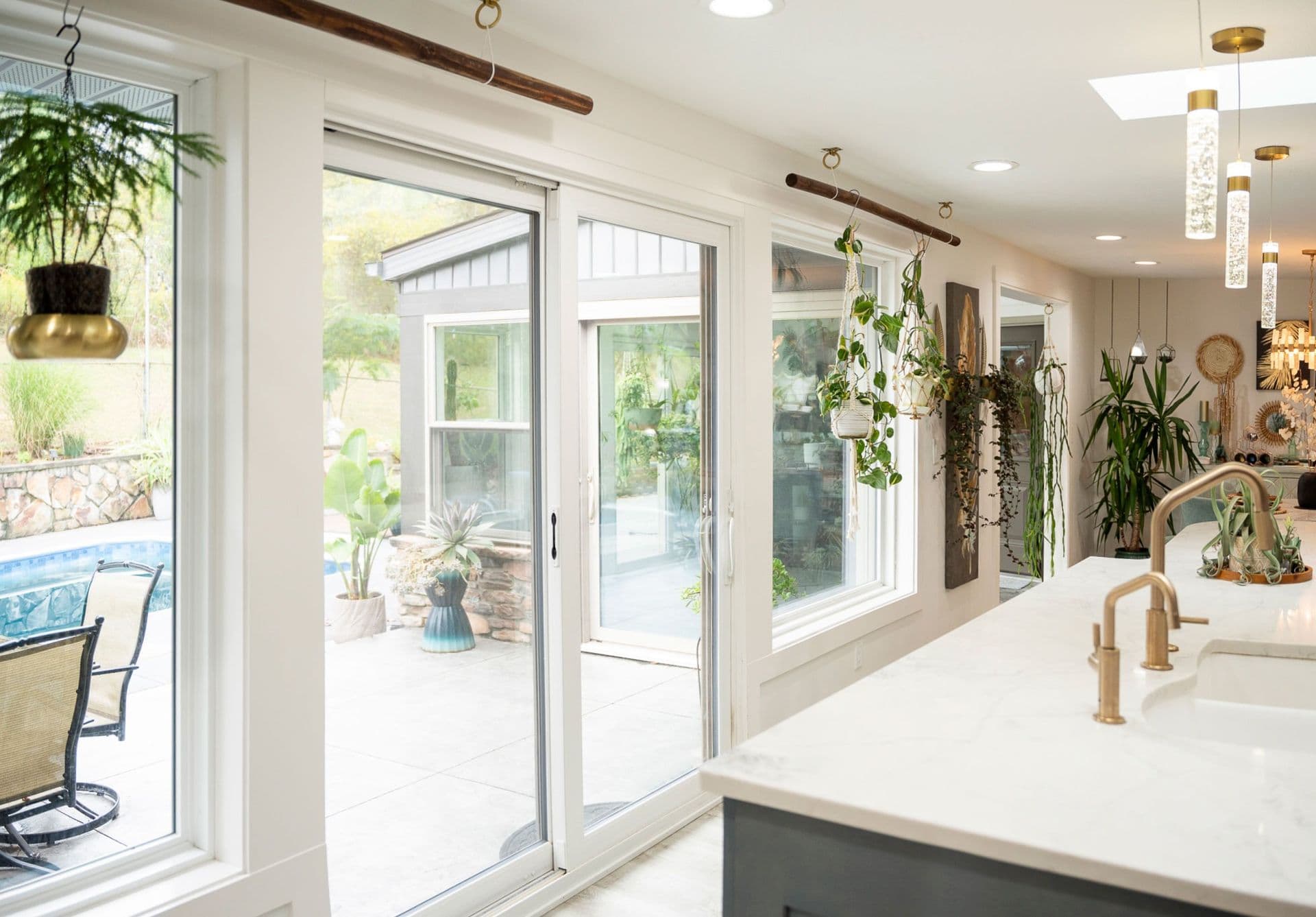 Interior view looking out through a white Endure 2-lite sliding glass patio door and surrounding picture windows toward a backyard pool patio. A modern kitchen island with a white marble countertop, brass gold faucets, pendant lights, and hanging plants occupies the foreground.