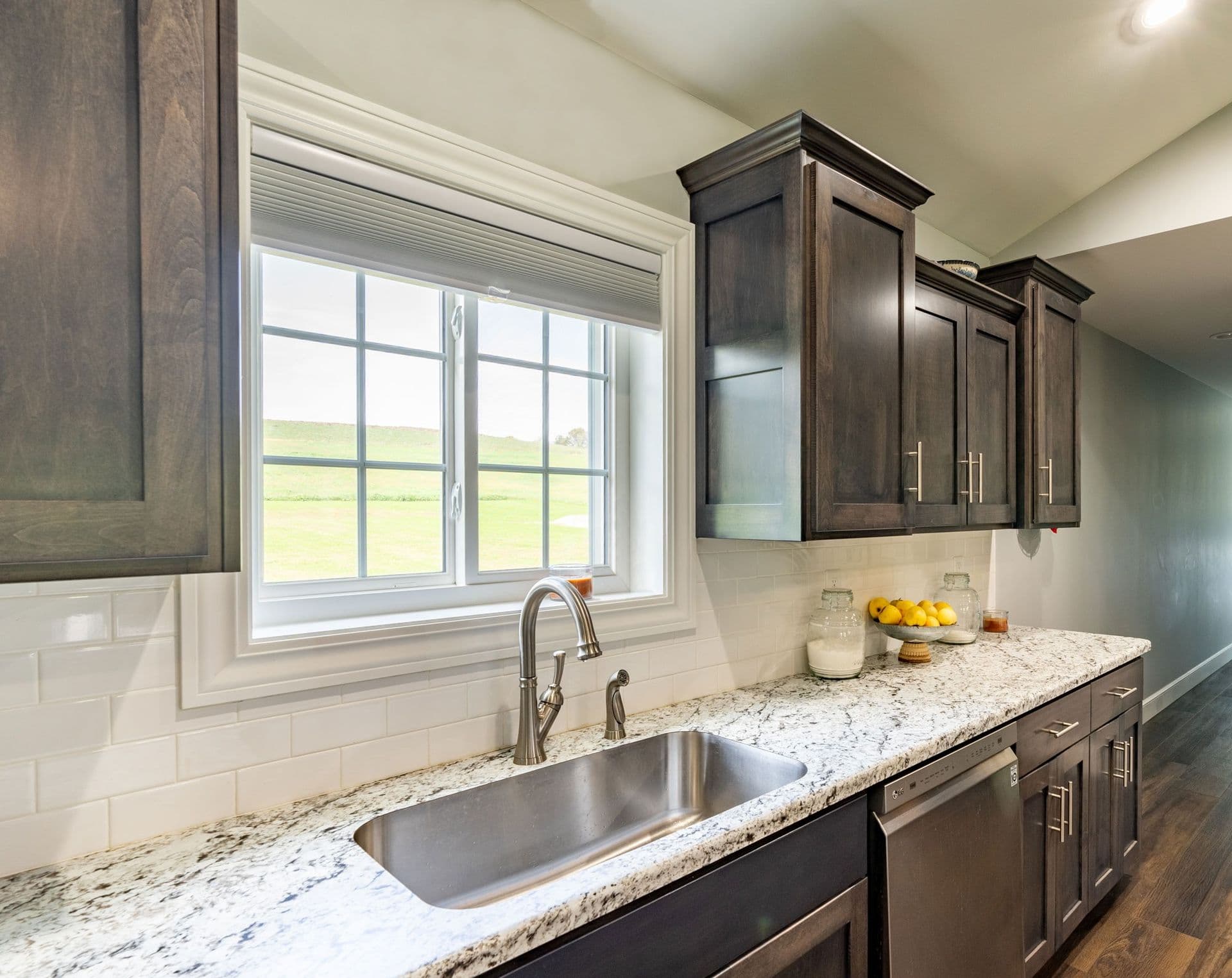 Interior kitchen view featuring a white slider window with grid panes installed above an undermount stainless steel sink with a gooseneck faucet. The countertop is granite with dark wood cabinetry and a white subway tile backsplash.