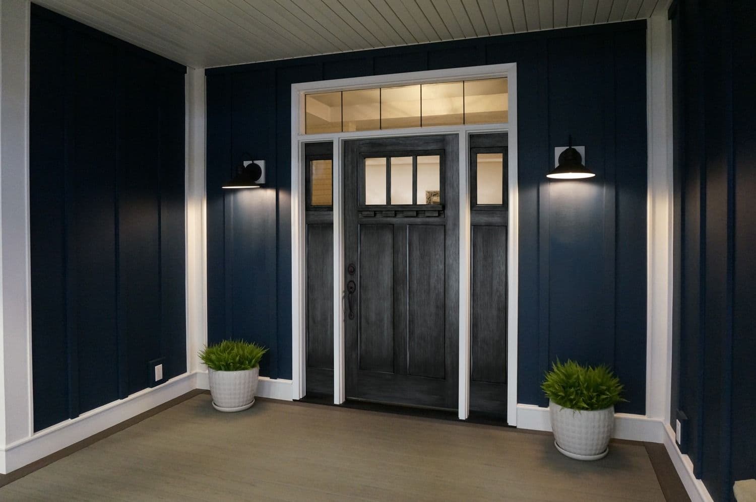 Exterior entryway view of a covered porch with deep navy blue vertical board-and-batten siding, featuring a dark charcoal Craftsman-style front door with four upper glass panes and matching sidelights set inside crisp white casing, lit by two black wall sconces and styled with potted green plants.