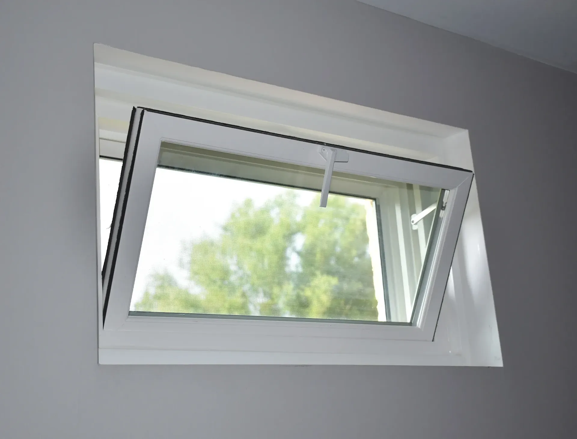 Interior view of a white-framed hopper window installed in a grey wall, hinged at the bottom and tilted inward from the top to allow ventilation.