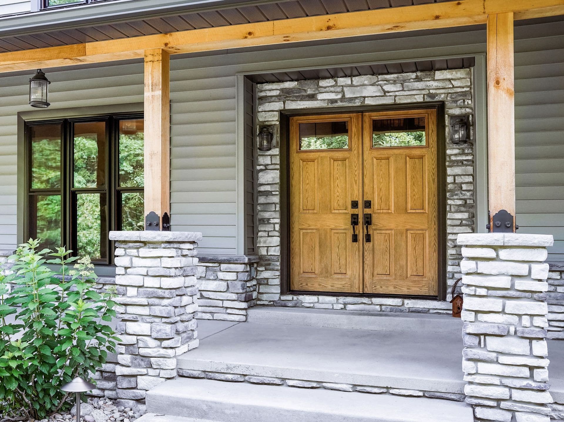 Exterior porch view featuring light oak-stained double entry doors with rectangular top glass windows, framed by light grey stacked stone veneer. The porch is supported by square timber posts with stone pedestals set against light grey lap siding and black-framed triple windows.