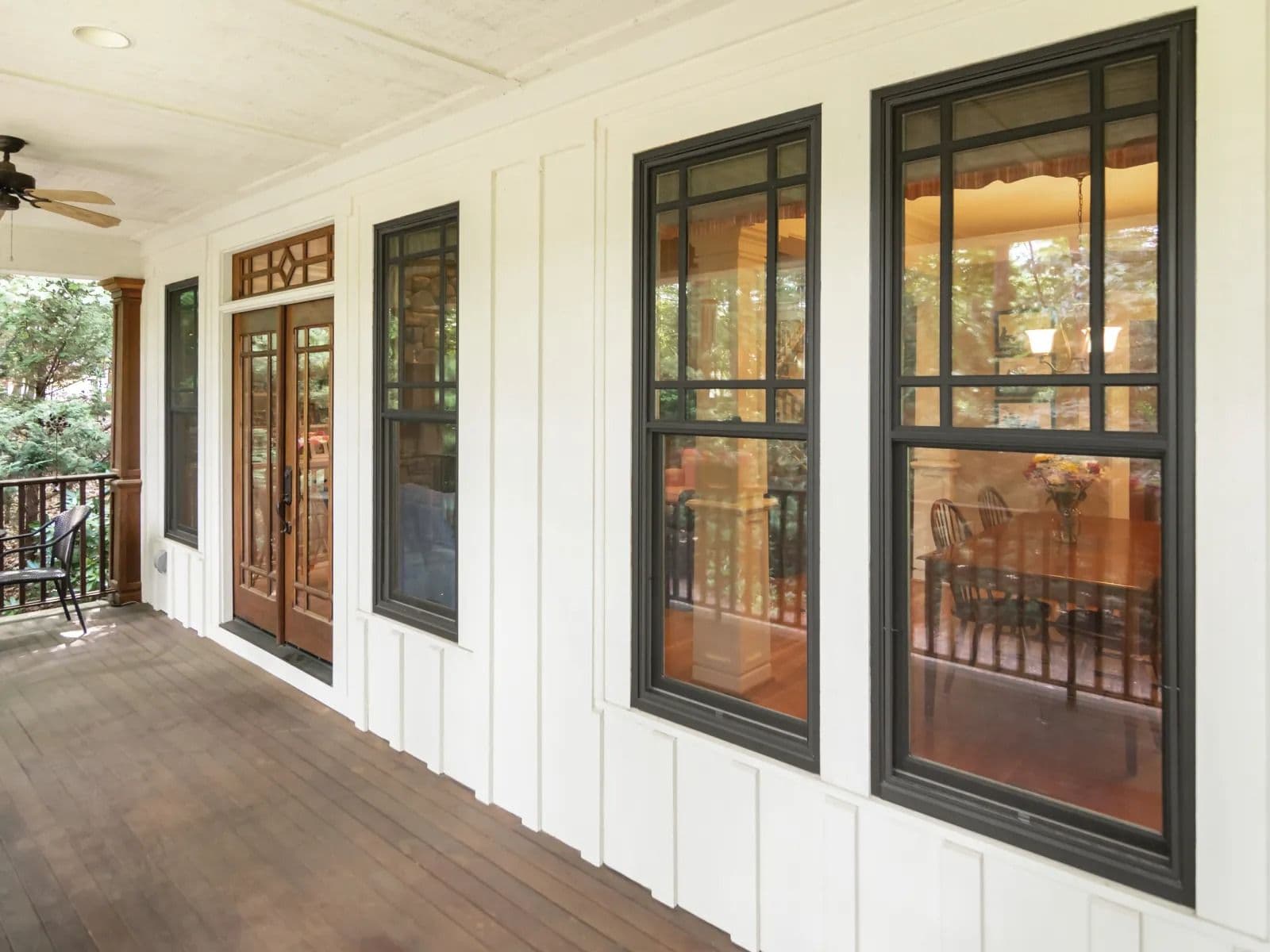 Exterior view of a covered wooden porch with white board-and-batten siding, featuring dark-framed double-hung windows with Prairie-style grids that reveal a dining room interior, alongside double glass patio doors and a ceiling fan overhead.