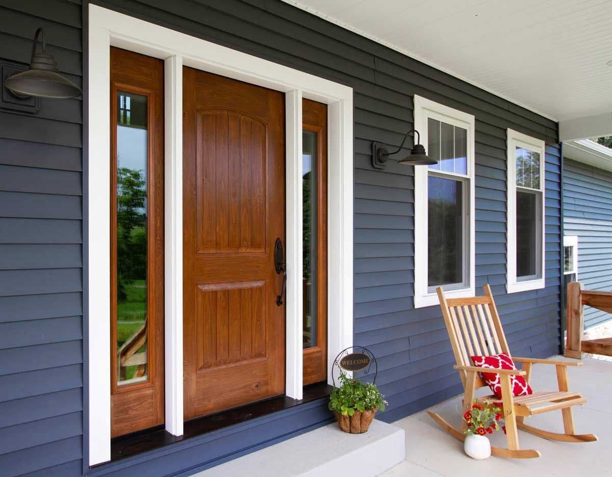 Exterior front porch view featuring a warm caramel-stained knotty alder entry door with two narrow glass sidelights set in white trim, against dark slate blue horizontal siding. The porch is furnished with a wooden rocking chair with a patterned red pillow, gooseneck wall sconces, and potted plants.