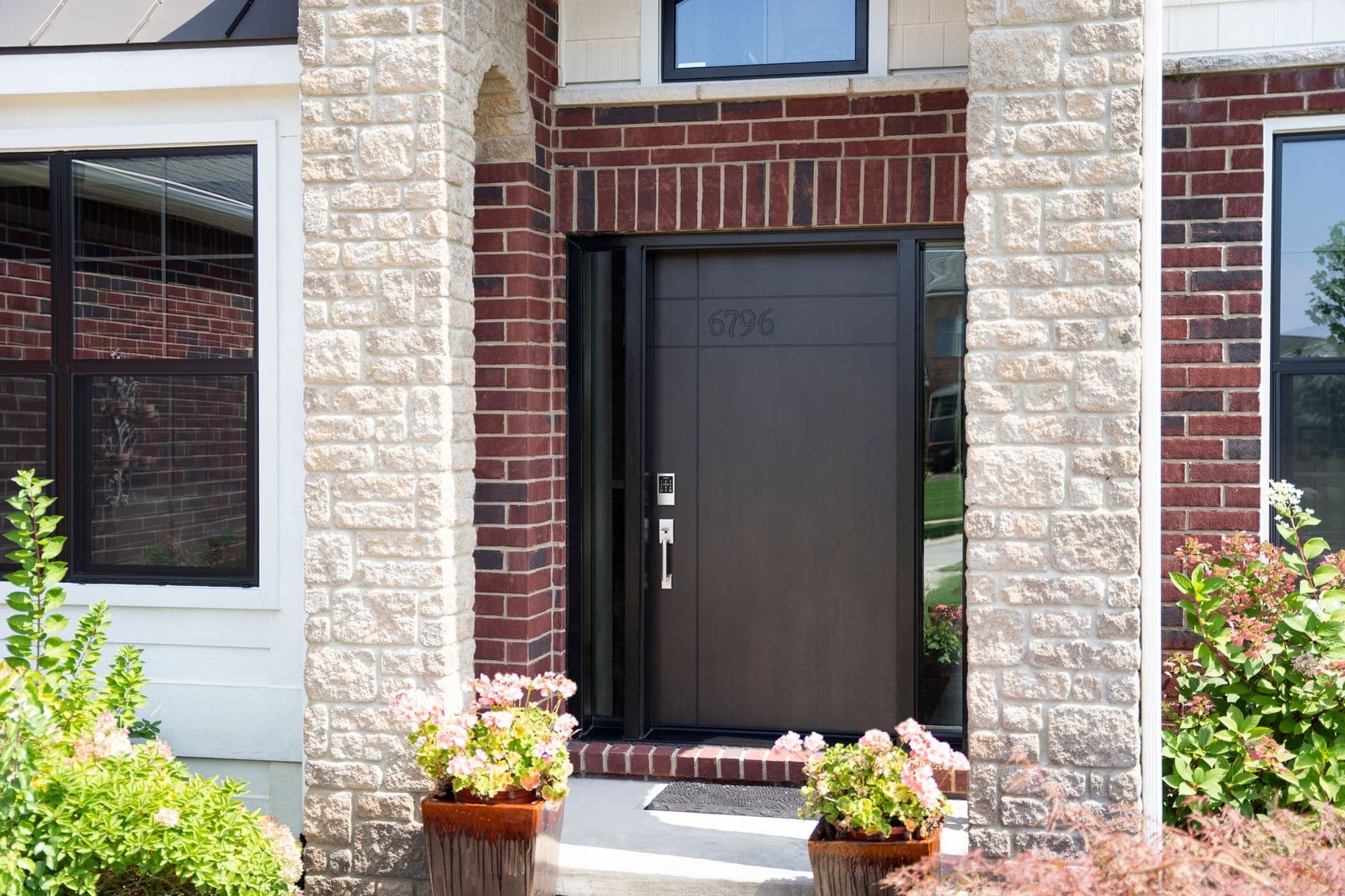 Exterior entryway view featuring a modern, dark espresso Coffee Bean-stained front door inscribed with the house number 6796 near the top, equipped with a digital keypad lock and modern silver handle set, flanked by full-length narrow glass sidelights and framed by light stone stone pillars, dark red brickwork, and potted pink flowers.