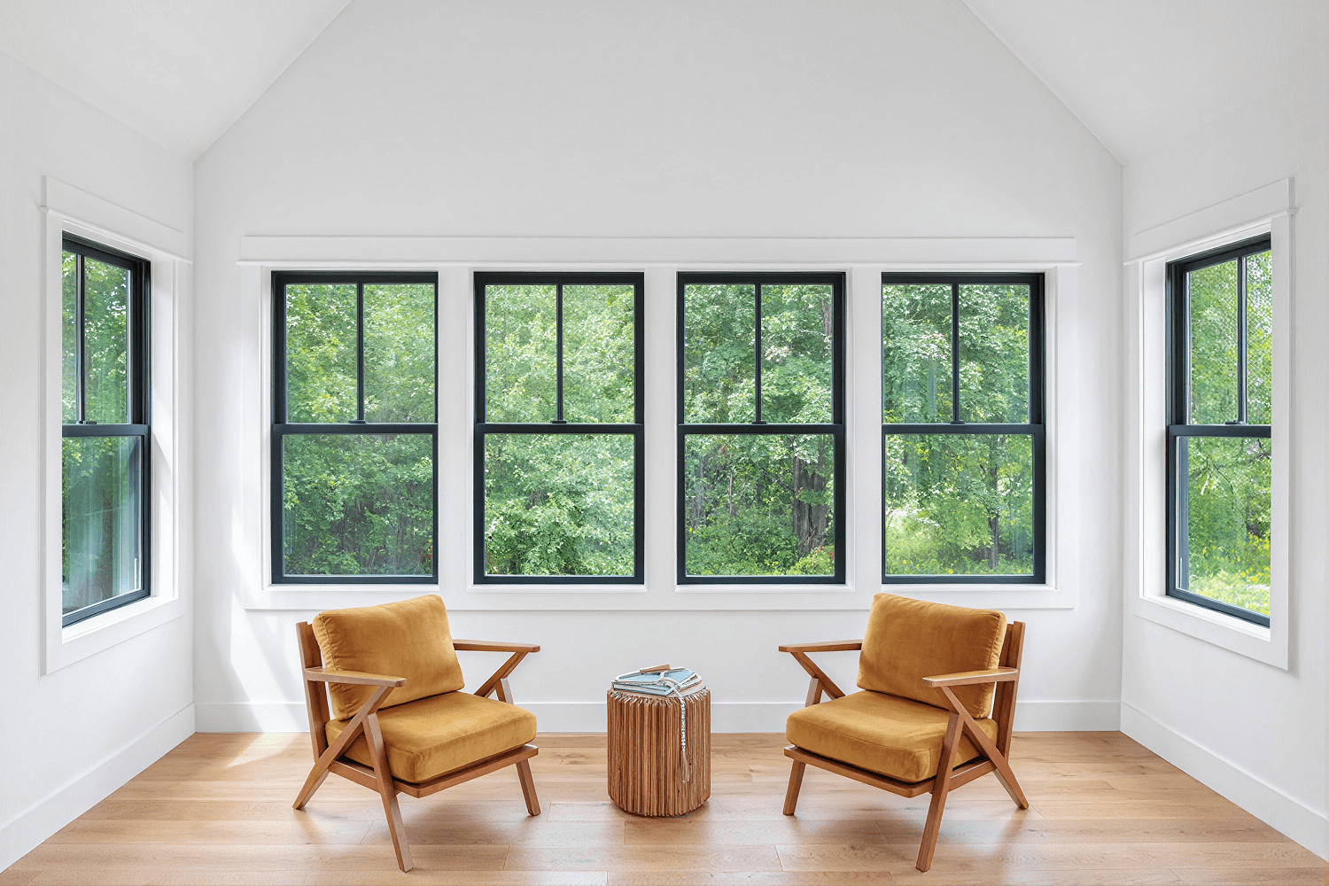 Interior view of a bright, sunlit room with vaulted white ceilings and light wood floors, featuring black double-hung windows along three walls that overlook a dense green forest. Two mustard-yellow upholstered armchairs with wooden frames face each other with a small round wooden accent table set between them.