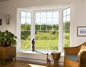 Interior view of a three-panel bay window projecting outward from a sunlit living room with white grid panes, offering a scenic view of a green field and trees.