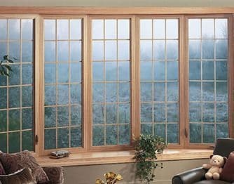 Interior view of a five-panel curved bow window with light wood frames and decorative muntin grids, looking out onto a misty landscape.