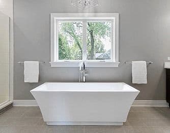 Interior view of a modern bathroom with a freestanding white bathtub positioned beneath a white-framed casement window. The room features grey walls, light grey tiled flooring, and white bath towels hanging on chrome wall racks on either side of the window.