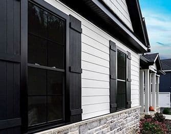 Exterior angle view of a modern home featuring black double-hung windows with dark grid panes and matching black decorative shutters. The building exterior combines white horizontal siding with a stacked stone veneer base along the foundation.