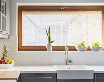 Interior kitchen view showing a wide hopper window with a warm wood frame installed above a white farmhouse sink and grey cabinets. Small potted plants sit along the wide window sill in front of white horizontal blinds.