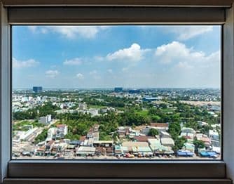 Interior view looking through a large, single-pane picture window that offers an expansive, unobstructed view of an urban landscape, lush green trees, and a partly cloudy blue sky.