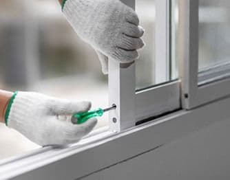 Close-up view of a technician wearing white work gloves using a green screwdriver to adjust or repair the frame mechanism of a white sliding glass window.