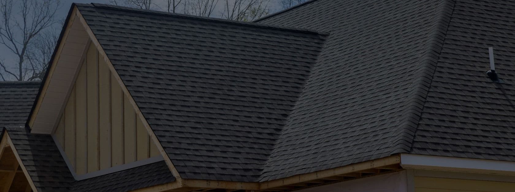 Exterior angle view of a residential roof featuring dark charcoal architectural asphalt shingles on multiple gables and roof planes, set against light tan vertical board-and-batten siding underneath the main peak.