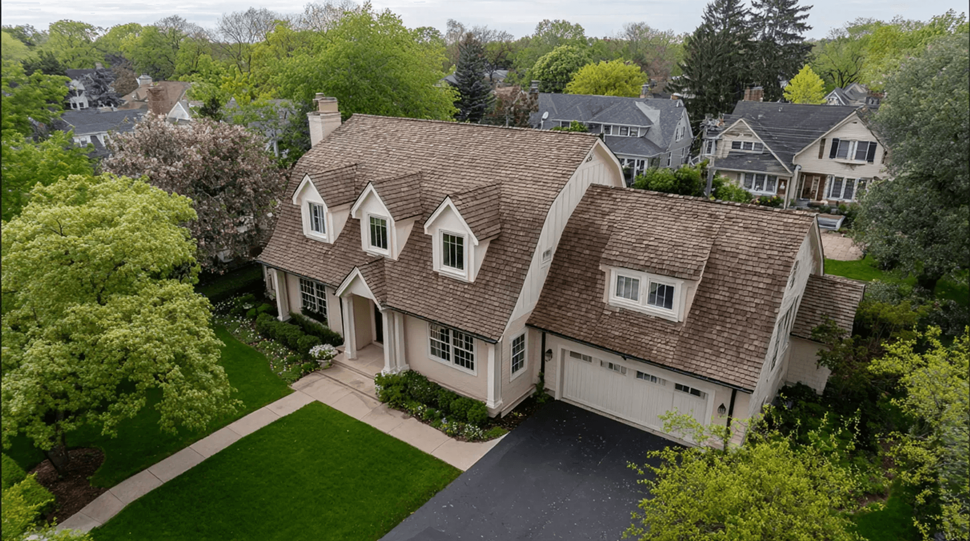 High-angle aerial view of a large two-story Cape Cod-style home featuring a brown cedar shake roof, off-white exterior, four dormer windows, an attached garage with a paved driveway, and a manicured green lawn surrounded by mature trees.