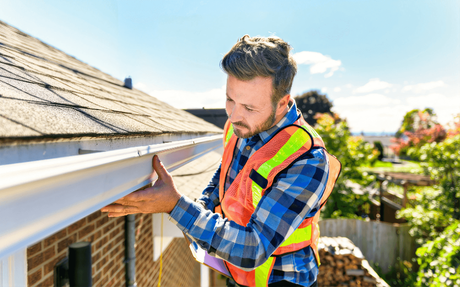 Close-up view of a home inspector wearing a bright safety vest and plaid shirt inspecting a white roof gutter system attached to the eave of a brick house.
