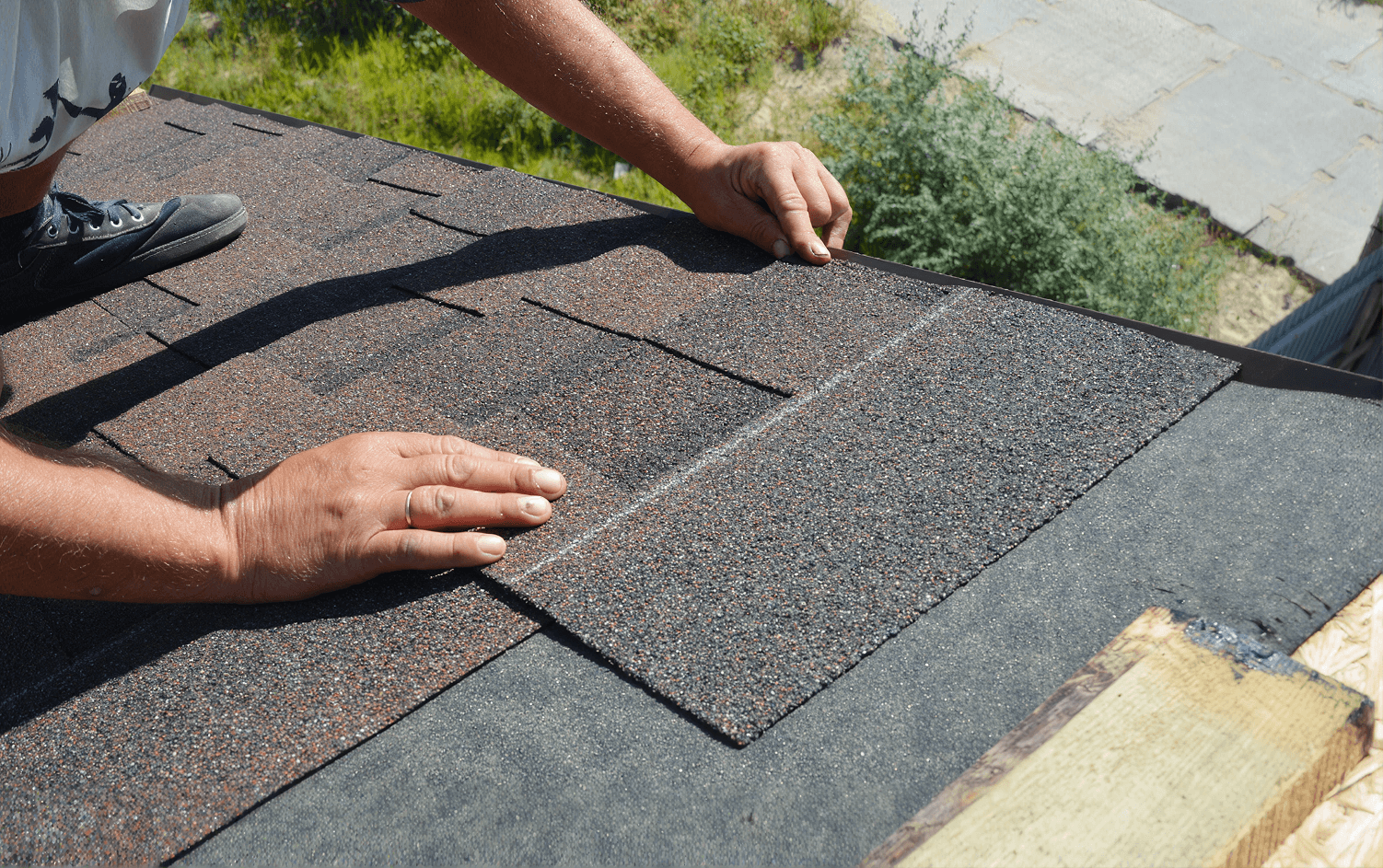 Close-up view of a roofer's hands carefully aligning and installing individual brown-and-grey asphalt shingles onto a rooftop surface covered with dark underlayment felt.